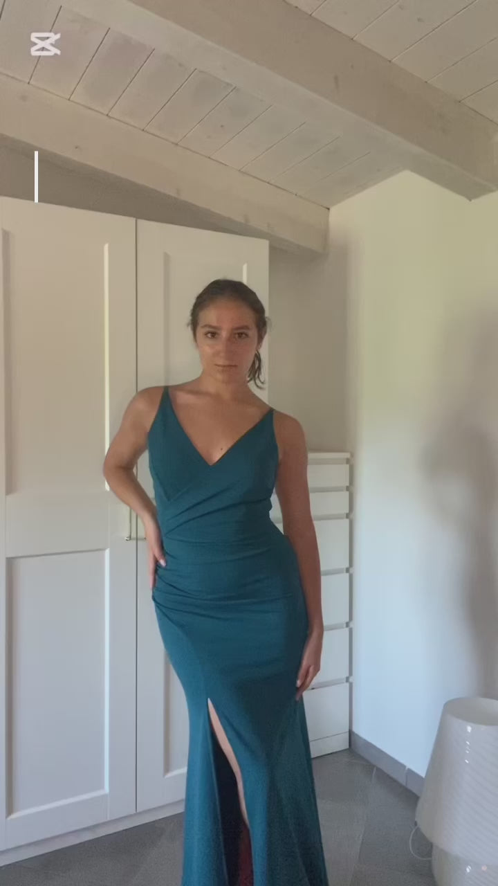 donna indossa un abito elegante blu scuro con spalline sottili e uno sguardo accattivante, posa in uno spoglio interno con armadio bianco sullo sfondo