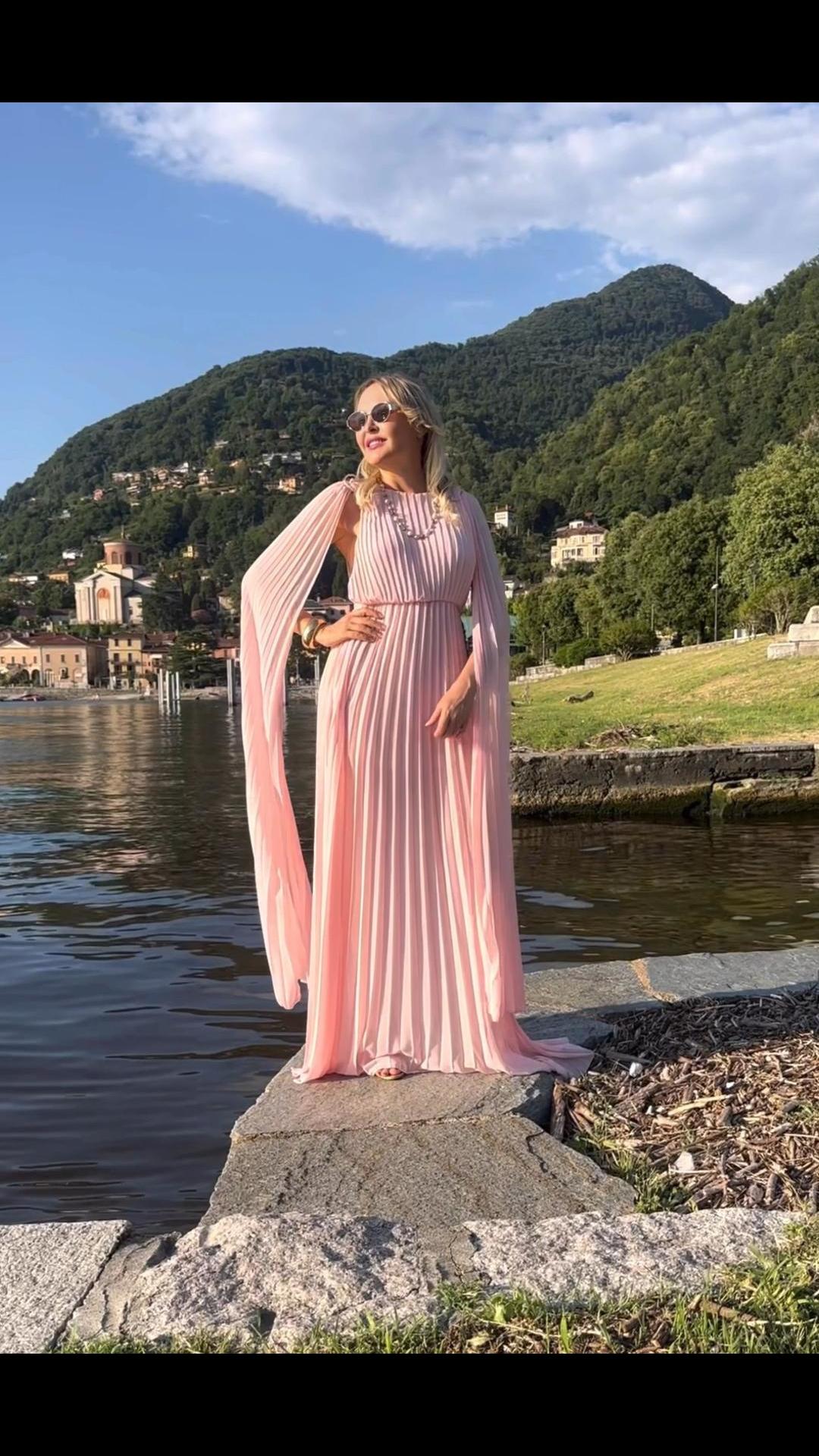 Donna con abito rosa plissé e mantelle, posa sul lago con montagne sullo sfondo