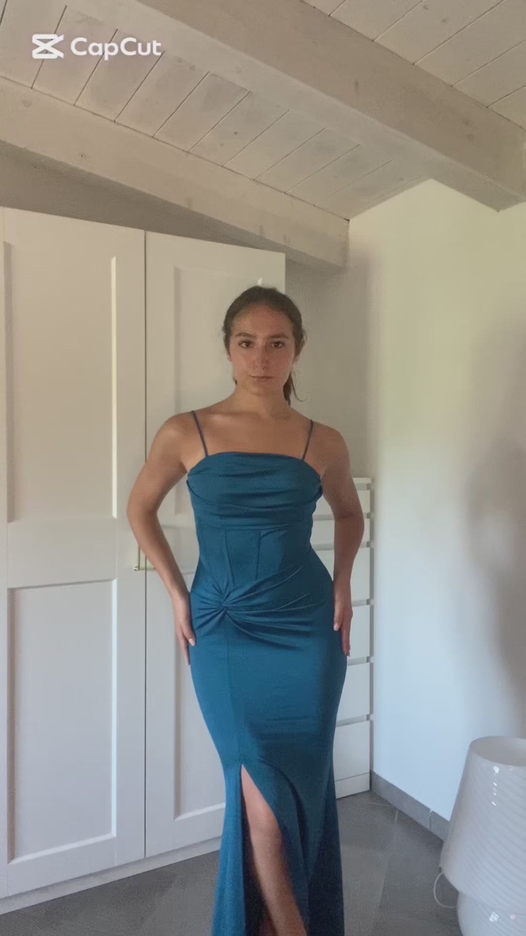 Una donna indossa un abito elegante blu scuro con spalline sottili e uno sguardo accattivante, posa in uno spoglio interno con armadio bianco sullo sfondo.