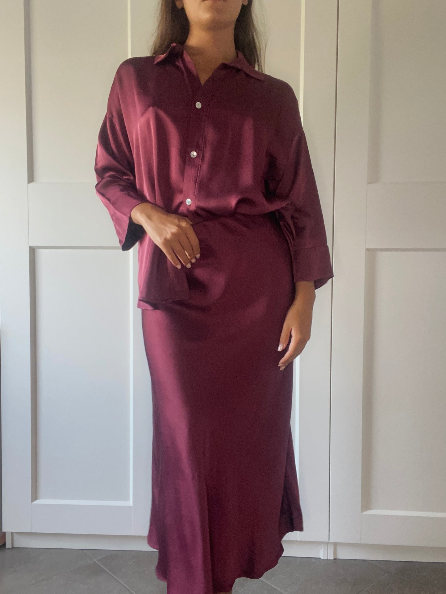Modella con un outfit elegante in seta bordeaux: camicia a bottoni e gonna lunga, in un ambiente domestico con porte bianche.