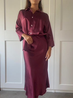 Modella con un outfit elegante in seta bordeaux: camicia a bottoni e gonna lunga, in un ambiente domestico con porte bianche.
