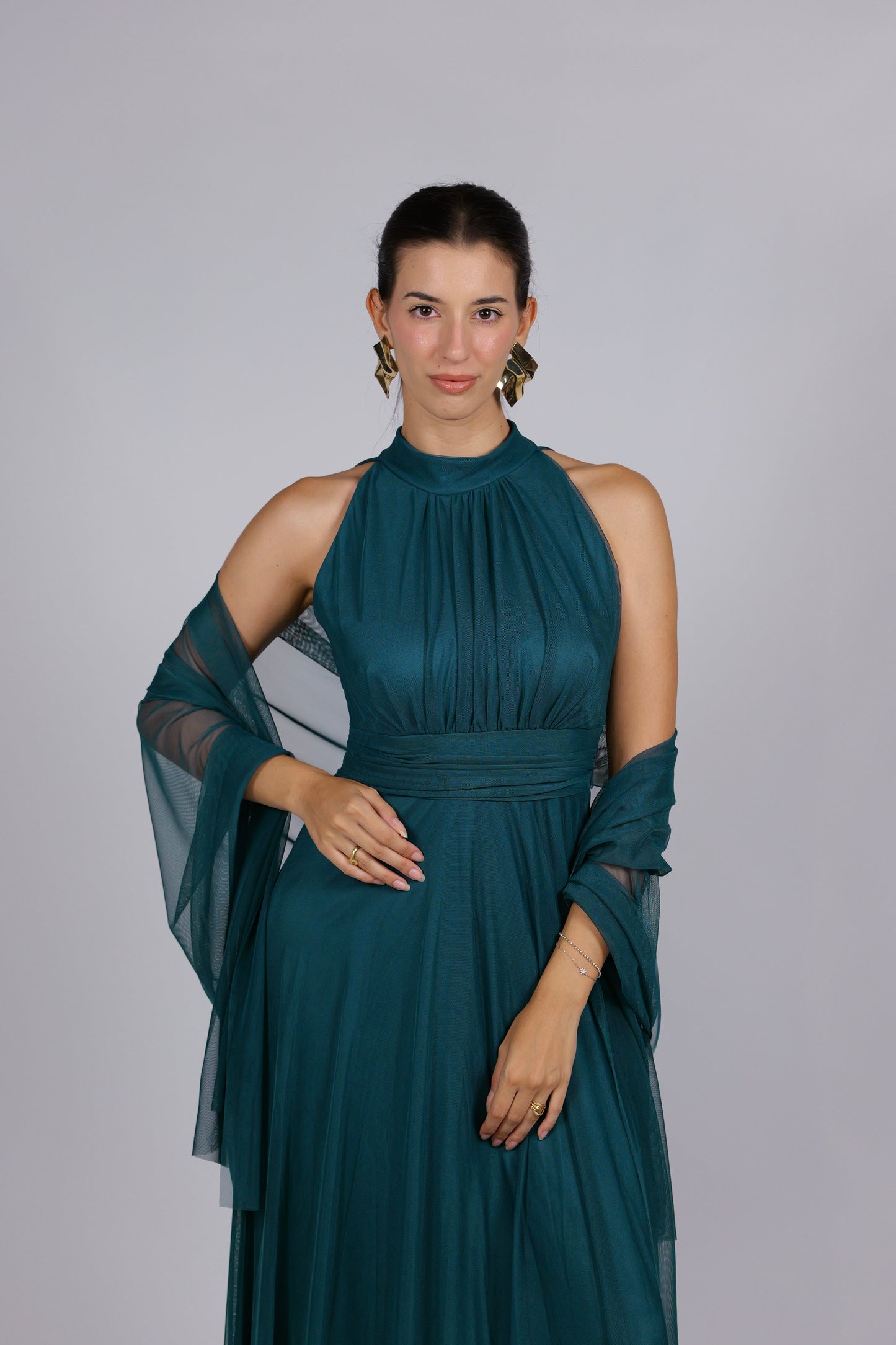 Donna con abito elegante di colore verde scuro, collo alto a imbarcatura, gonna lunga, con foulard in tulle trasparente, su sfondo chiaro.
