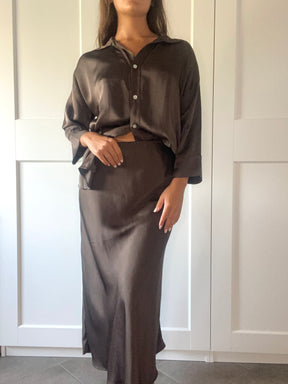 Donna indossa un camicione e una gonna in seta fango, entrambi a lunghezza massima, posando in un ambiente chiaro.