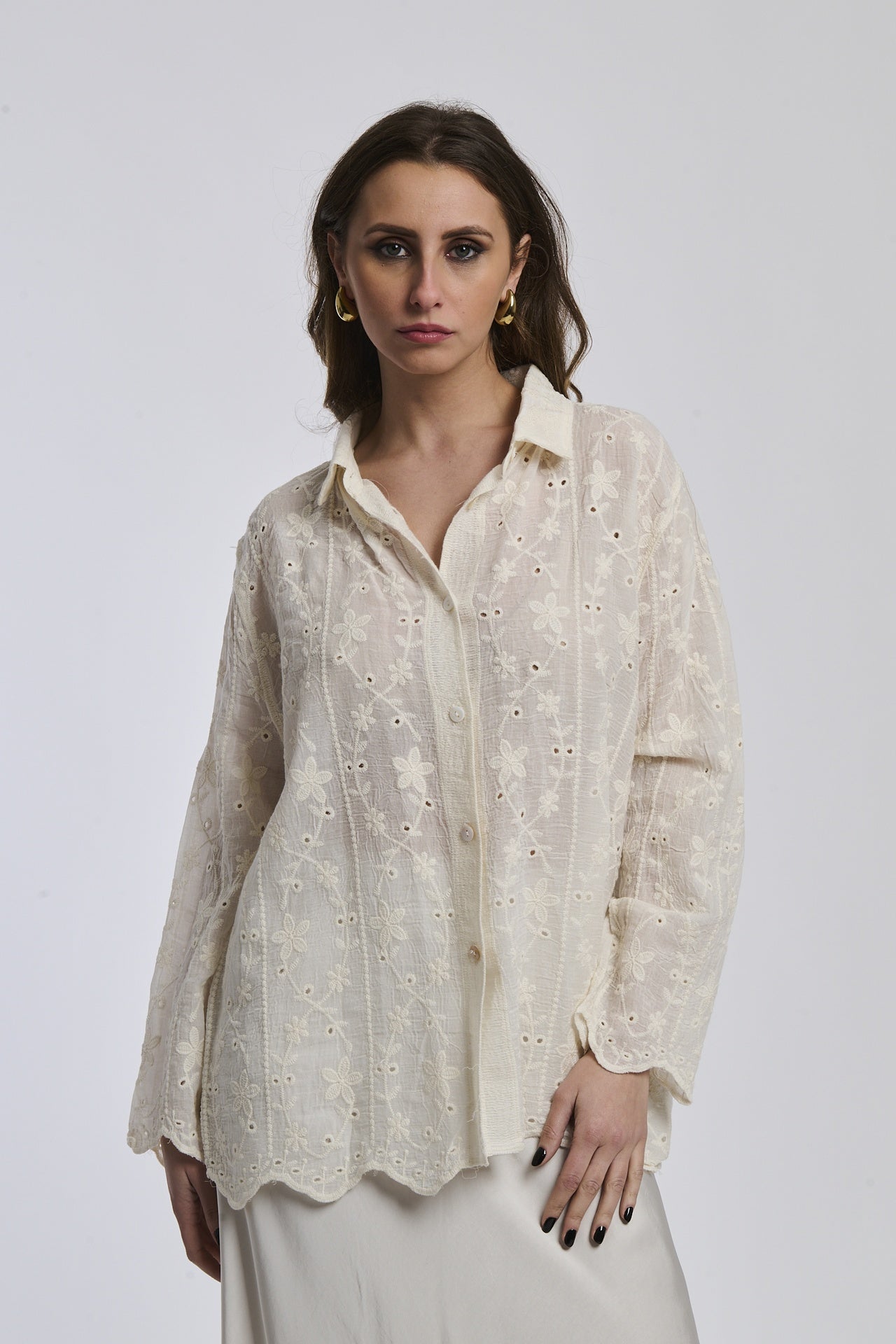 Donna con camicia bianca ricamata, colletto a bottone e orecchini dorati, sfondo beige
