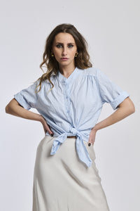 Donna con camicia a righe azzurre, colletto a imbuto e nodo frontale, che conferisce un’eleganza leggera.