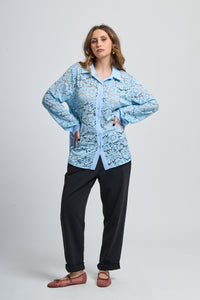 Camicetta azzurra con motivi, collo camiciera e maniche lunghe, abbinata a pantaloni neri.