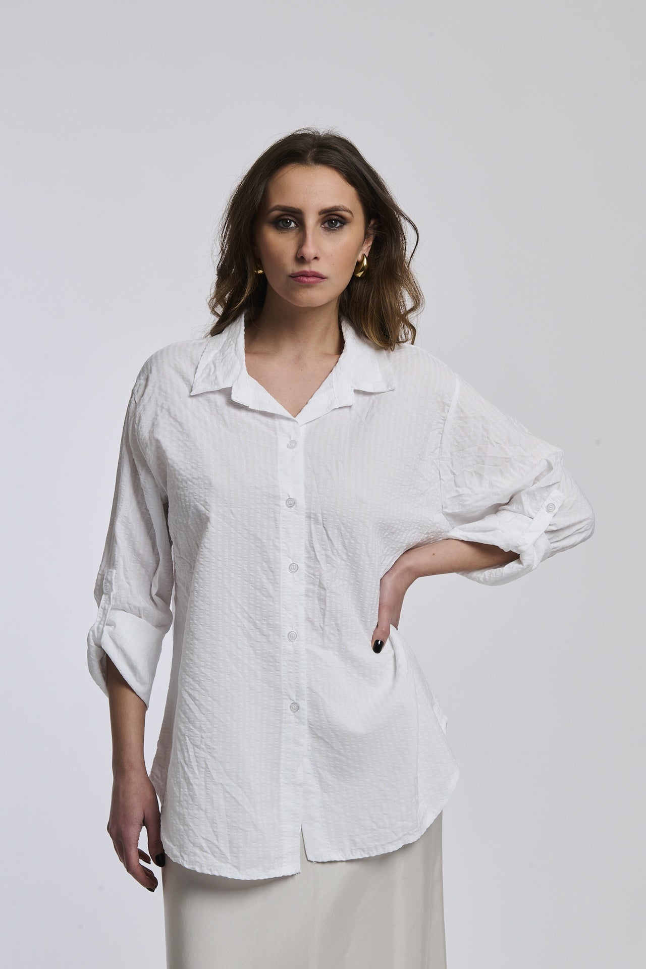Donna indossa una camicia a righe bianche, oversize, colletto a camicia, bottoni frontali e maniche arrotolate.