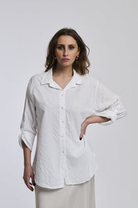 Donna indossa una camicia a righe bianche, oversize, colletto a camicia, bottoni frontali e maniche arrotolate.