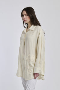 Camicia di un colore beige e intenso indossata dalla donna, con maniche arrotolate e un leggero slittamento, che trasmette un senso di mistero e profondità emotiva.