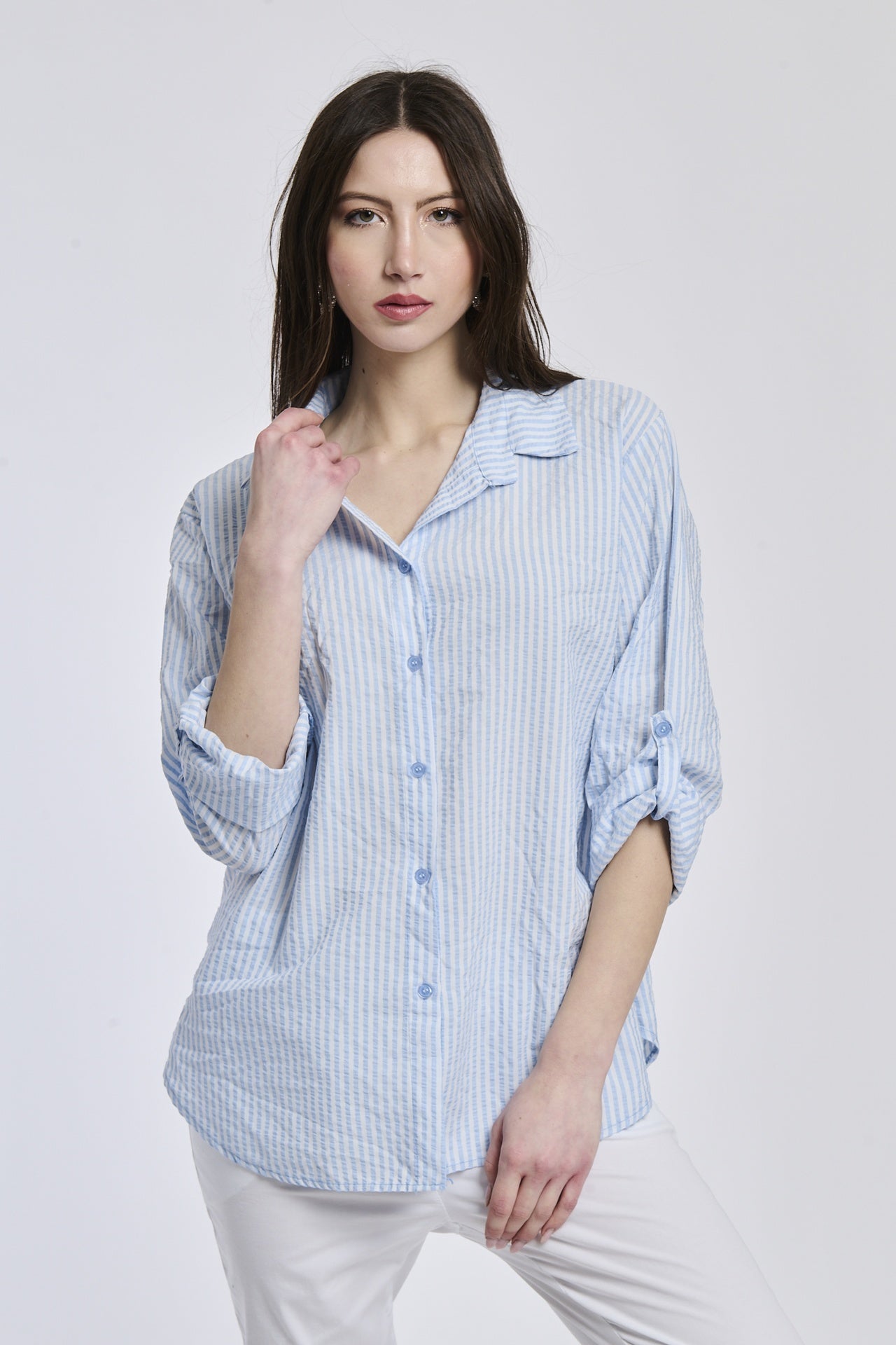 Donna indossa una camicia a righe azzurre, oversize, colletto a camicia, bottoni frontali e maniche arrotolate.
